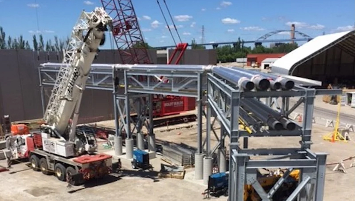 A large white construction crane lifting heavy piping onto an elevated metal support structure at an outdoor industrial job site.